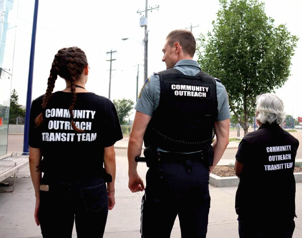 Three adults of various ages stand shoulder to shoulder wearing vests that say "community outreach" on the back.