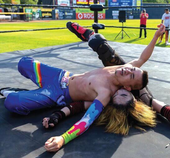 A person in rainbow clothing tackles another wrestler in the ring