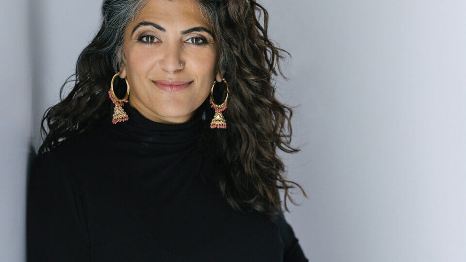 An photo of a South Asian woman with a white background. The woman has long, black hair. She is wearing a black turtleneck and beaded hoop earrings.
