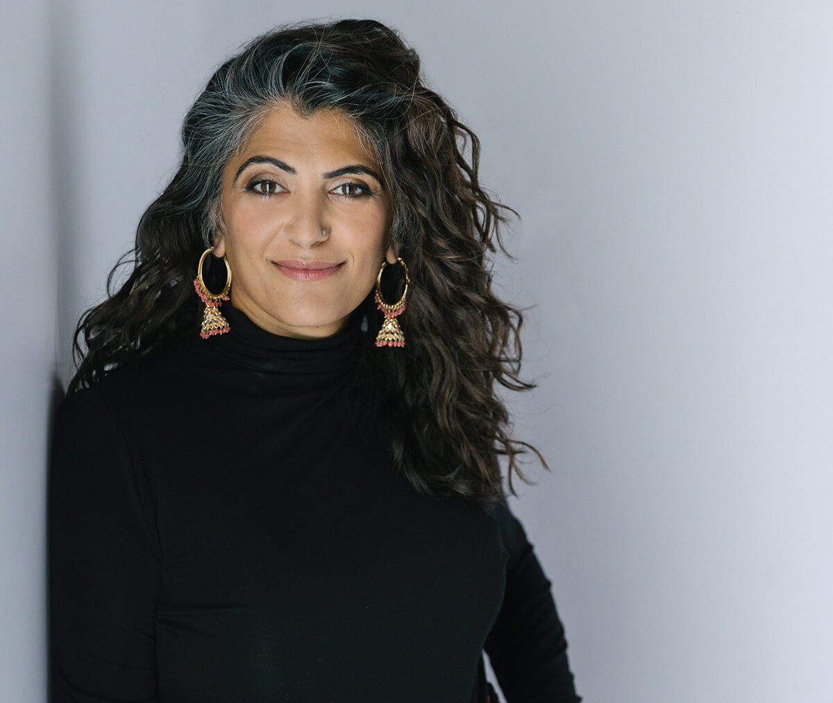 An photo of a South Asian woman with a white background. The woman has long, black hair. She is wearing a black turtleneck and beaded hoop earrings.