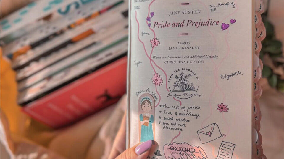 A photo of a hand holding up the inside cover page of Pride and Prejudice. It has been annotated with doodles. A bag full of books is out of focus in the background.