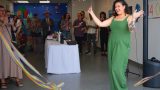 Photo of a small gathering with people standing around a table with a white cloth. In the foreground, rainbow streamers wave into the image, and a woman in a green dress stands and smiles with her arms held out in celebration.