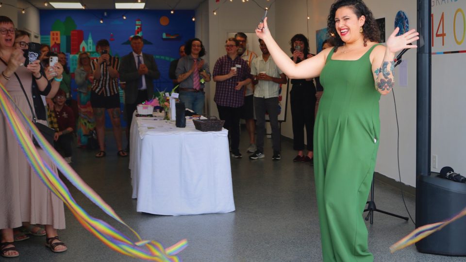 Photo of a small gathering with people standing around a table with a white cloth. In the foreground, rainbow streamers wave into the image, and a woman in a green dress stands and smiles with her arms held out in celebration.