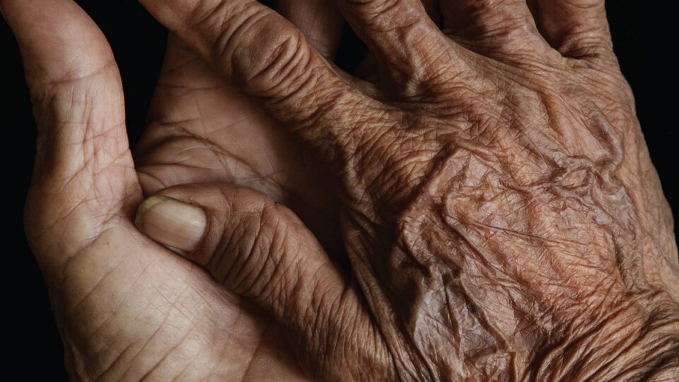Close-up photo of wrinkled, brown hands against a black background.