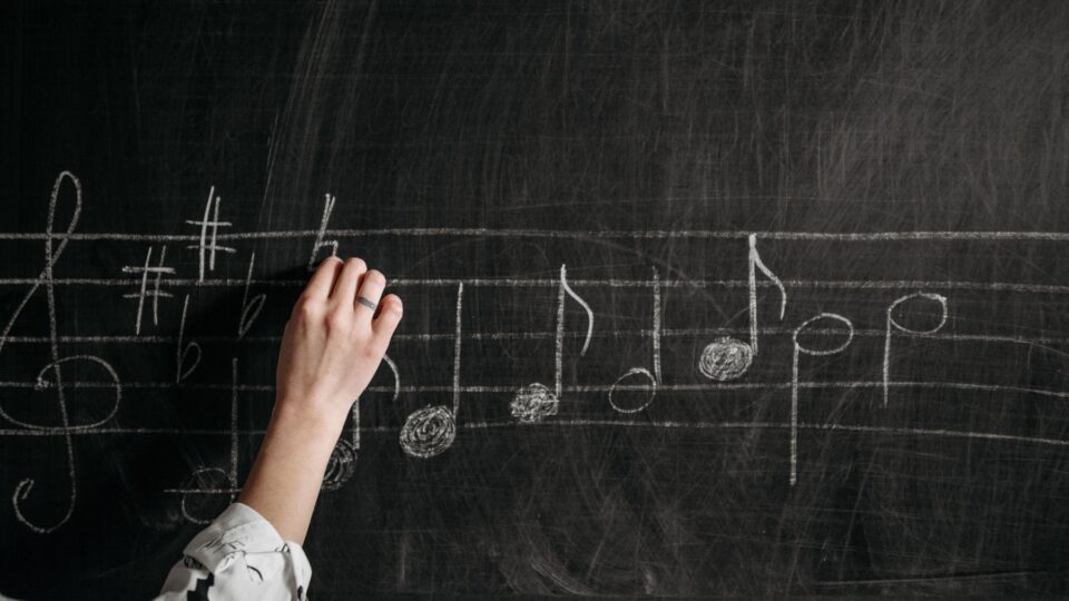Photo of a hand writing music notes in white chalk on a dusty blackboard.
