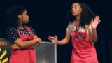 Photo of two black women wearing red aprons acting on a stage.