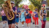 Photo of members of a marching band playing their instruments and walking down a tree-lined street.