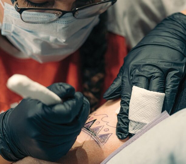 Photo of a tattoo artist in a mask and glasses tattooing a design onto a person with a handpoke tool.
