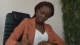 Photo of a Black woman with braids wearing an orange blazer and sitting behind a desk writing.